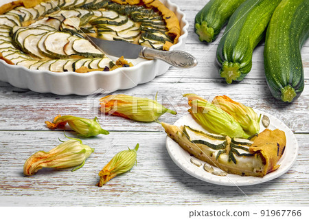 Open pie of zucchini and cream cheese filling on a white plate and in a baking dish surrounded by flowers Open pie of zucchini and cream cheese filling on a white plate and in a baking dish surrounded by flowers 91967766