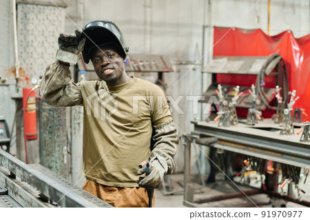Portrait of African young welder in mask smiling at camera while working with metal products at factory Portrait of African young welder in mask smiling at camera while working with metal products at factory 91970977