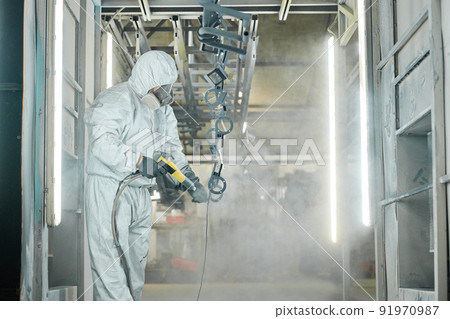 Man in protective suit spraying powder paint from gun on metal products at factory Man in protective suit spraying powder paint from gun on metal products at factory 91970987