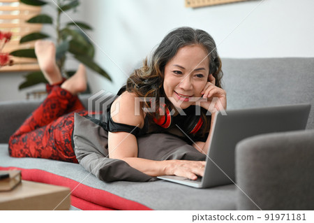 Smiling middle aged woman chatting online in social network, watching movie on laptop while lying on couch 91971181