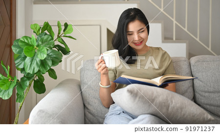 Pretty young asian woman sitting on couch and reading interesting book, enjoying leisure weekend time at home Pretty young asian woman sitting on couch and reading interesting book, enjoying leisure weekend time at home 91971237