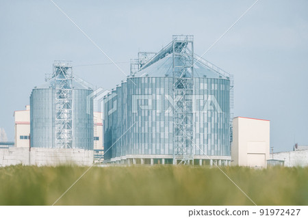 Grain elevator. Metal grain elevator in agricultural zone. Agriculture storage for harvest. Grain elevators on green nature background. Exterior of agricultural factory. Nobody. Grain elevator. Metal grain elevator in agricultural zone. Agriculture storage for harvest. Grain elevators on green nature background. Exterior of agricultural factory. Nobody. 91972427