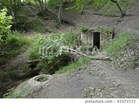 Mysterious stone door in the middle of the forest. Dark portal to the hill among old rocks. Broken trees and moss around. Sunny day. High quality photo. Mysterious stone door in the middle of the forest. Dark portal to the hill among old rocks. Broken trees and moss around. Sunny day. High quality photo. 91973572
