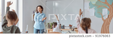 Young happy elementary school pupils at light modern classroom are rising their hands on a lesson 91976063