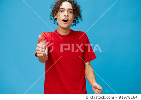 Excited shocked funny curly tanned Caucasian guy in basic red t-shirt show thumb up gesture Like posing isolated on blue background. Lifestyle and Emotions concept. Good offer with copy space for ad 91979164