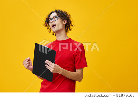 Joyful happy myopic young student man in red t-shirt funny eyewear holds tablet folder with study notes posing isolated on over yellow studio background. Free place for ad. Education College concept Joyful happy myopic young student man in red t-shirt funny eyewear holds tablet folder with study notes posing isolated on over yellow studio background. Free place for ad. Education College concept 91979165