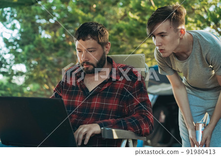 Caucasian bearded man and teenage son working wing with laptop outdoors. Camping concept Caucasian bearded man and teenage son working wing with laptop outdoors. Camping concept 91980413