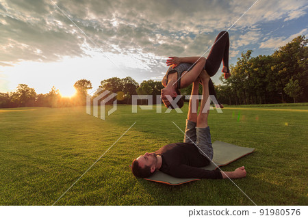 Man lying on grass and balancing woman in his feet. Young couple doing acro yoga in park at sunset 91980576