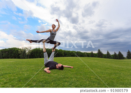 Man lying on grass and balancing woman in his feet. Young couple doing acro yoga in park 91980579