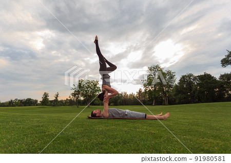 Healthy man lying on grass and balancing woman. Couple doing acrobatic yoga exercise in park 91980581