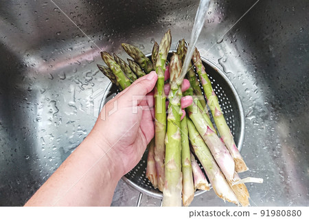 Womans hand wash fresh green asparagus sprouts in strainer in kitchen sink, personal perspective view. 91980880