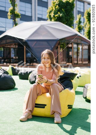 A beautiful young woman is sitting in a binbag outdoors in the city and writing a message on her smartphone A beautiful young woman is sitting in a binbag outdoors in the city and writing a message on her smartphone 91980985