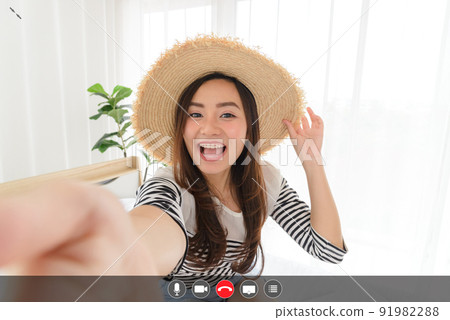 Young Asian woman blogger smiling in summer casual clothes and straw hat talking to facetime video calling with smartphone in bedroom. Using videochat online app with mic, video, phone, share screen, 91982288