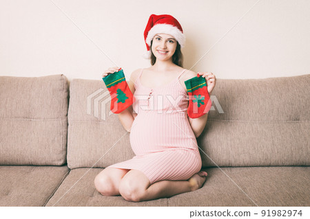 Cute happy pregnant woman in dress at home in santa hat. Mother to be with big belly, hands over tummy. Merry Christmas holidays. New Year Eve concept Cute happy pregnant woman in dress at home in santa hat. Mother to be with big belly, hands over tummy. Merry Christmas holidays. New Year Eve concept 91982974