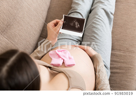 Pregnant Woman sitting on bed and Looking At Ultrasound Scan photo of her Baby. A pregnant female shows her ultrasound report and her unborn baby 91983019