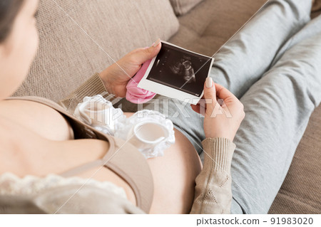 Pregnant Woman sitting on bed and Looking At Ultrasound Scan photo of her Baby. A pregnant female shows her ultrasound report and her unborn baby Pregnant Woman sitting on bed and Looking At Ultrasound Scan photo of her Baby. A pregnant female shows her ultrasound report and her unborn baby 91983020