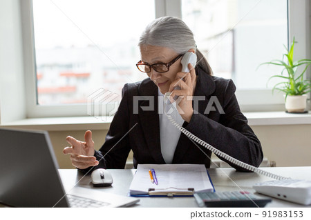 Attractive caucasian elderly woman in formal wear and eyeglasses sitting at the desk in office, talks on a stationary phone and gesturing with her hand, selective focus. 91983153