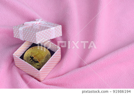 The golden bitcoin lies in a small pink gift box with a small bow on a blanket made of soft and fluffy light pink fleece fabric with a large number of relief folds The golden bitcoin lies in a small pink gift box with a small bow on a blanket made of soft and fluffy light pink fleece fabric with a large number of relief folds 91986194