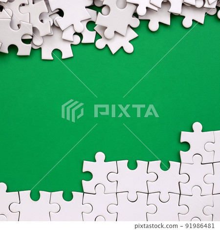 Fragment of a folded white jigsaw puzzle and a pile of uncombed puzzle elements against the background of a green surface. Texture photo with space for text Fragment of a folded white jigsaw puzzle and a pile of uncombed puzzle elements against the background of a green surface. Texture photo with space for text 91986481