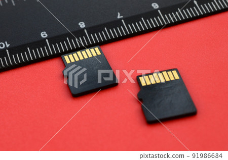 Two small micro SD memory cards lie on a red background next to a black ruler. A small and compact data and information store Two small micro SD memory cards lie on a red background next to a black ruler. A small and compact data and information store 91986684