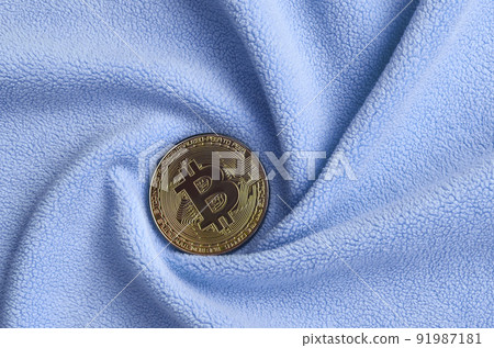 The golden bitcoin lies on a blanket made of soft and fluffy light blue fleece fabric with a large number of relief folds. The shape of the folds resembles a fan from a video card cooler The golden bitcoin lies on a blanket made of soft and fluffy light blue fleece fabric with a large number of relief folds. The shape of the folds resembles a fan from a video card cooler 91987181