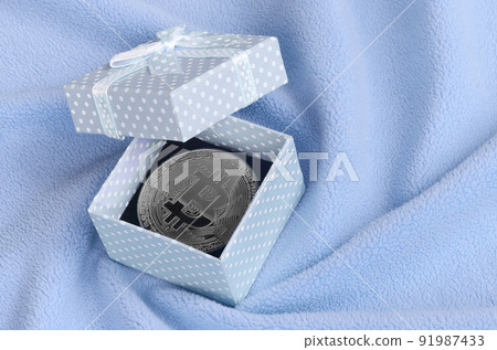 The silver bitcoin lies in a small blue gift box with a small bow on a blanket made of soft and fluffy light blue fleece fabric with a large number of relief folds The silver bitcoin lies in a small blue gift box with a small bow on a blanket made of soft and fluffy light blue fleece fabric with a large number of relief folds 91987433