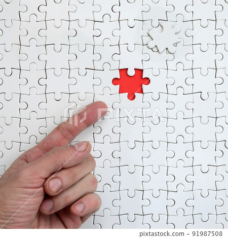 The texture of a white jigsaw puzzle in the assembled state with one missing element, forming a red space, pointed to by the finger of the male hand 91987508