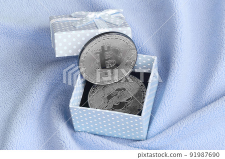 The silver bitcoin lies in a small blue gift box with a small bow on a blanket made of soft and fluffy light blue fleece fabric with a large number of relief folds The silver bitcoin lies in a small blue gift box with a small bow on a blanket made of soft and fluffy light blue fleece fabric with a large number of relief folds 91987690