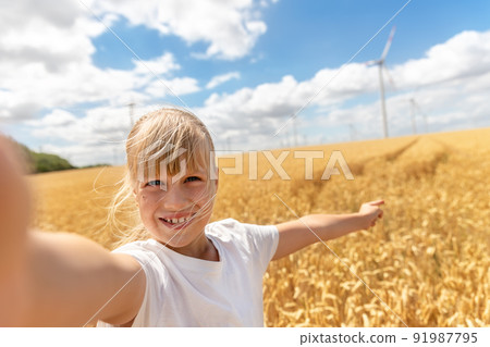 Portrait of little cute beautiful blond caucasian girl enjoy having fun taking selfie photo phone camera walking at wheat field against wind mill turbine farm. Children blogging and video streaming Portrait of little cute beautiful blond caucasian girl enjoy having fun taking selfie photo phone camera walking at wheat field against wind mill turbine farm. Children blogging and video streaming 91987795