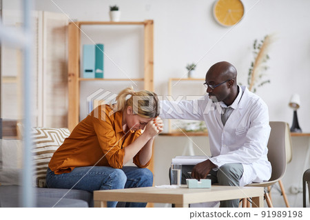 Understanding young Black psychiatrist in white coat and glasses touching shoulder of crying female patient while consoling her at therapy session 91989198