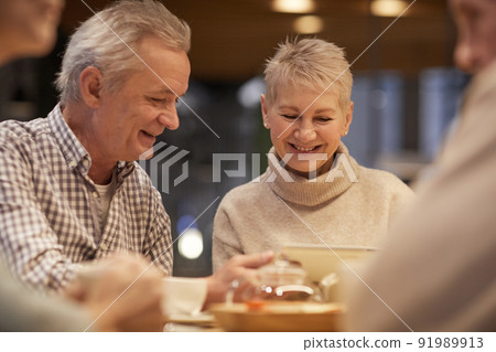 Cheerful modern seniors in casual outfits sitting in cafe and using tablet while discussing social media accounts 91989913