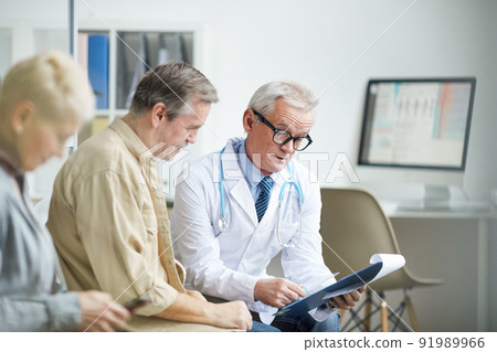 Serious aged male doctor with stethoscope around neck reading medical history of patient while holding consultation Serious aged male doctor with stethoscope around neck reading medical history of patient while holding consultation 91989966