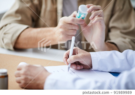 Close-up of unrecognizable male doctor sitting at table and filling prescription while patient reading pill bottle label Close-up of unrecognizable male doctor sitting at table and filling prescription while patient reading pill bottle label 91989989