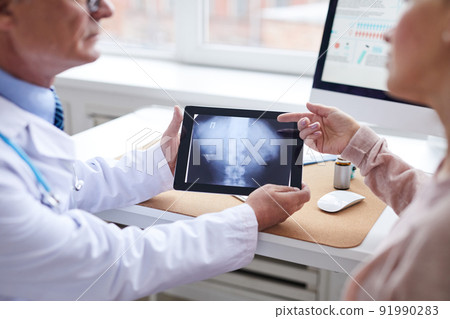 Close-up of female patient sitting table and pointing at tablet screen while asking question about online spine x-ray image to doctor 91990283