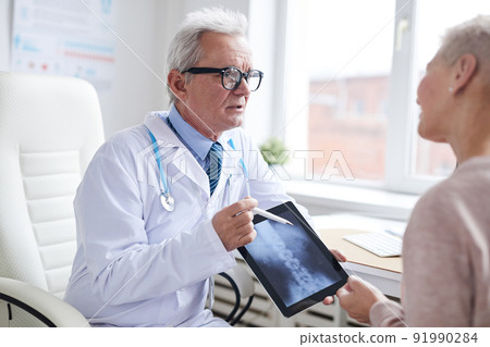 Confident senior male doctor in lab coat using digital tablet while telling patient about spinal cord disorder 91990284