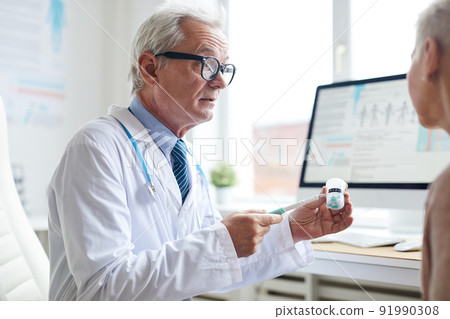 Serious wrinkled physician in glasses pointing with pen at pill bottle while advising patient to take medicine Serious wrinkled physician in glasses pointing with pen at pill bottle while advising patient to take medicine 91990308