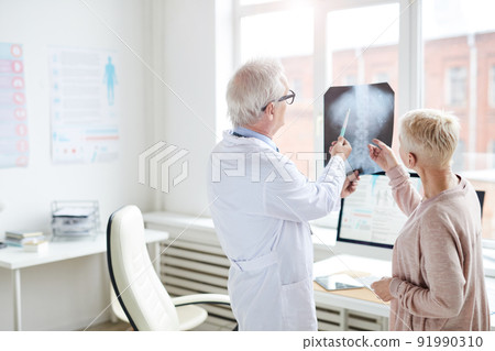 Rear view of gray-haired doctor in lab coat standing against window and watching x-ray image of spinal cord with female patient Rear view of gray-haired doctor in lab coat standing against window and watching x-ray image of spinal cord with female patient 91990310