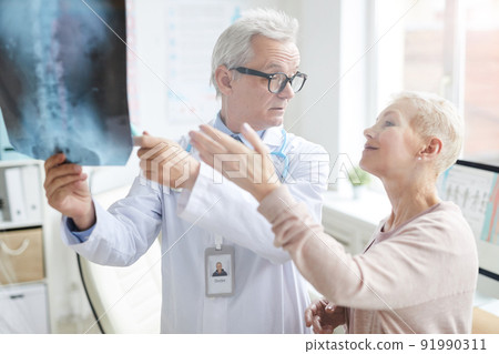 Attractive blond-haired mature lady pointing at core x-ray image while discussing her pain with doctor 91990311