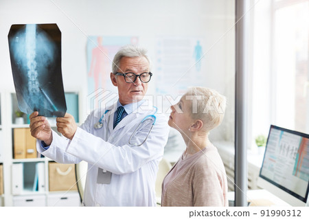 Frowning handsome aged doctor in eyeglasses holding x-ray image against light while explaining spinal problems to senior female patient Frowning handsome aged doctor in eyeglasses holding x-ray image against light while explaining spinal problems to senior female patient 91990312