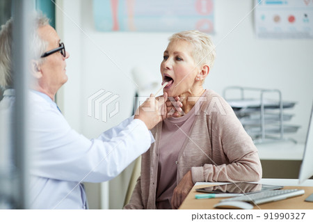 Serious aged male doctor in lab coat using tongue depressor while checking throat of mature lady during medical exam Serious aged male doctor in lab coat using tongue depressor while checking throat of mature lady during medical exam 91990327