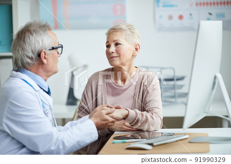 Senior doctor in eyeglasses sitting at table and holding hand of mature female patient while giving hope and comfort to her Senior doctor in eyeglasses sitting at table and holding hand of mature female patient while giving hope and comfort to her 91990329