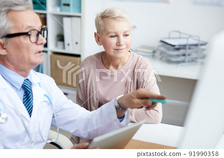 Serious senior male doctor sitting at table and pointing at monitor while explaining CAT scan to senior patient 91990358