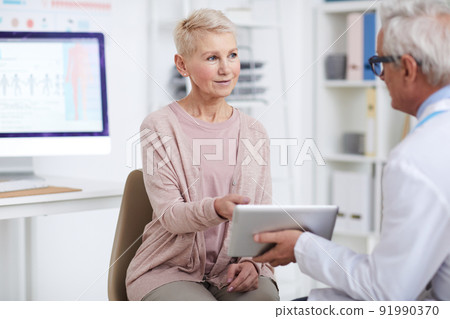 Content elderly woman in casual outfit sitting on chair and asking doctor about prescribed pills presented on tablet screen Content elderly woman in casual outfit sitting on chair and asking doctor about prescribed pills presented on tablet screen 91990370