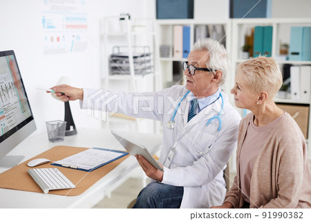 Confident senior male practitioner with gray hair sitting at table and pointing at computer screen while consulting mature female patient in clinic Confident senior male practitioner with gray hair sitting at table and pointing at computer screen while consulting mature female patient in clinic 91990382