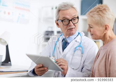 Serious wrinkled senior doctor in lab coat sitting at table and using digital tablet with online patients records while giving recommendations to mature woman Serious wrinkled senior doctor in lab coat sitting at table and using digital tablet with online patients records while giving recommendations to mature woman 91990383
