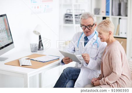 Content senior male doctor with badge and stethoscope sitting at table and using tablet while explaining medical checkups result to mature woman 91990393