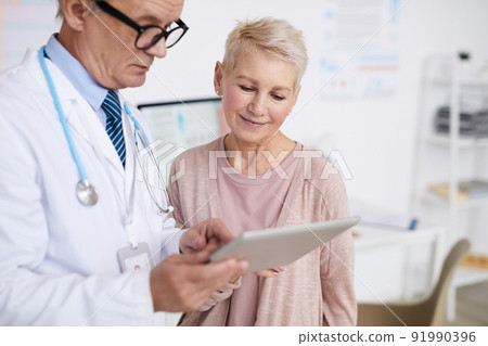 Senior practitioner with stethoscope around neck standing in clinic room and answering patients questions according to test results on tablet Senior practitioner with stethoscope around neck standing in clinic room and answering patients questions according to test results on tablet 91990396