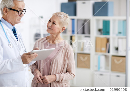 Smiling attractive mature blond-haired women in cardigan standing in doctors office and communicating with practitioner who using tablet Smiling attractive mature blond-haired women in cardigan standing in doctors office and communicating with practitioner who using tablet 91990408