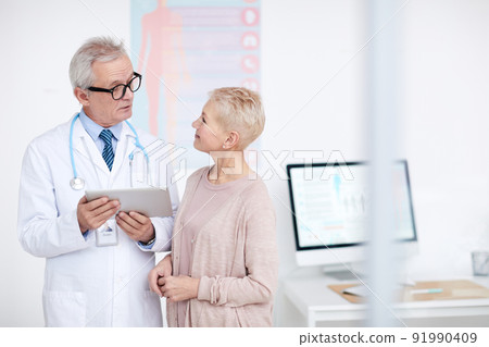 High-qualified senior gray-haired doctor in eyeglasses and lab coat standing in clinic room and counseling mature female patient High-qualified senior gray-haired doctor in eyeglasses and lab coat standing in clinic room and counseling mature female patient 91990409