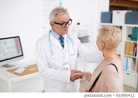 Serious frowning senior doctor in eyeglasses and white coat standing in own office and shaking hand of patient while talking to her Serious frowning senior doctor in eyeglasses and white coat standing in own office and shaking hand of patient while talking to her 91990412